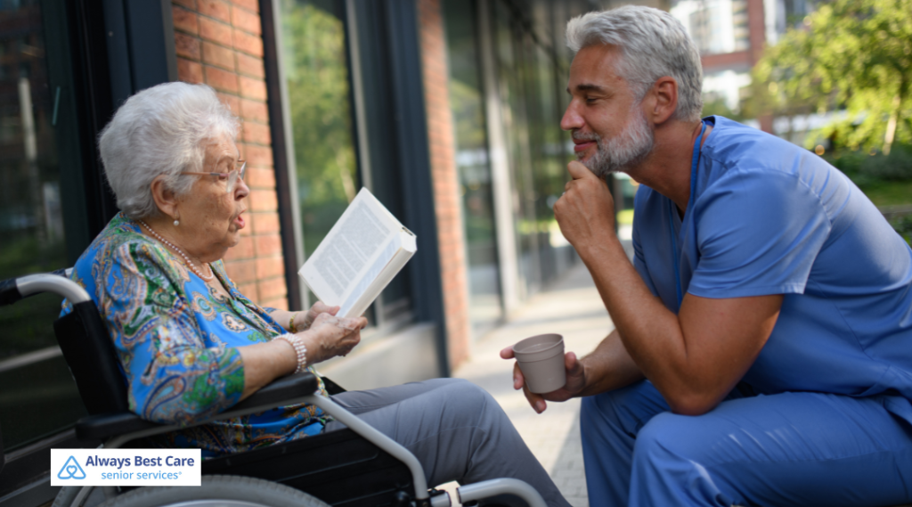 An elderly woman sits outside in a wheelchair, reading a book, while a male caregiver in blue scrubs kneels nearby holding a coffee cup and attentively engages with her. 