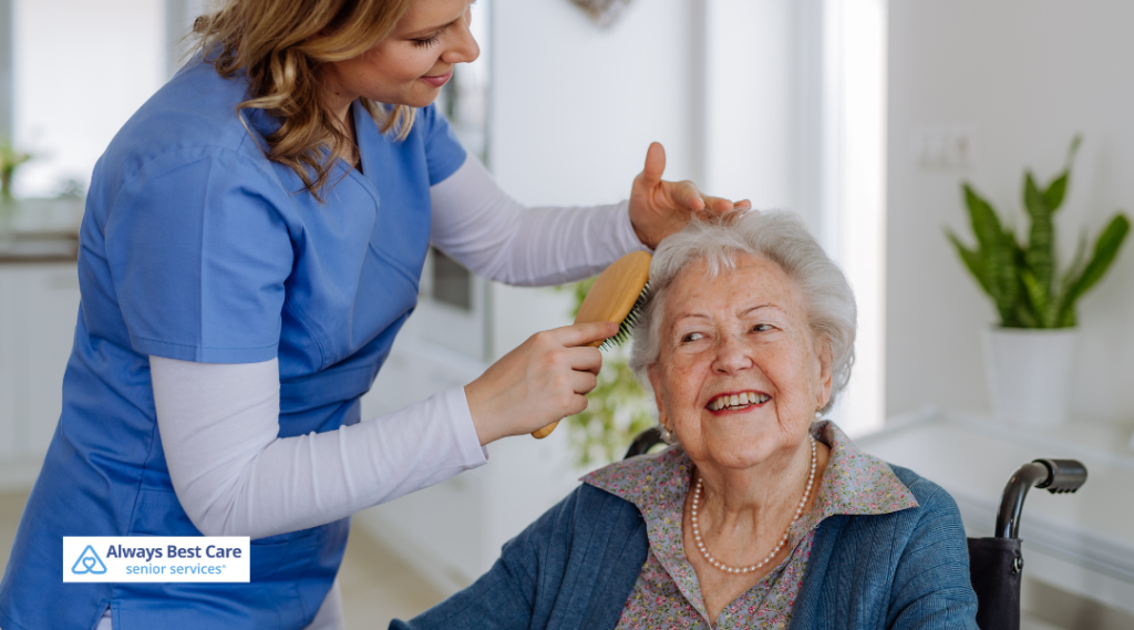 A caregiver in blue scrubs gently brushes the hair of a senior woman seated in a wheelchair, providing personal care in a well-lit and neatly decorated home environment. 