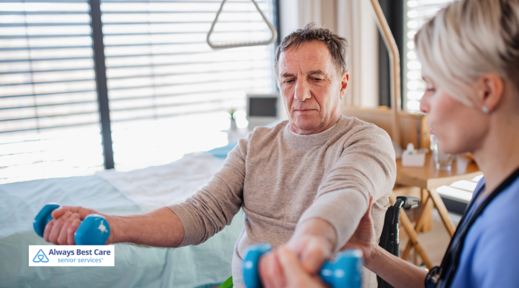 A man in a wheelchair performs arm exercises using light blue dumbbells while being guided by a female caregiver, as part of physical therapy in a comfortable bedroom setting.