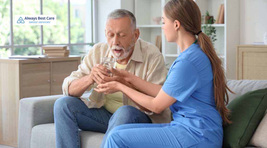 Caregiver in blue scrubs helps an older man hold a glass of water while he sits on a couch in a bright living room; ‘Always Best Care senior services’ logo in the corner.