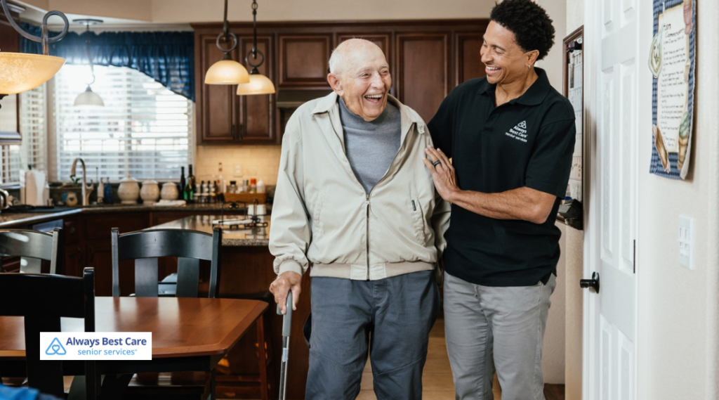 Caregiver in a black polo walks beside an older man using a cane in a kitchen/dining area; both smiling; ‘Always Best Care senior services’ logo in the corner.”