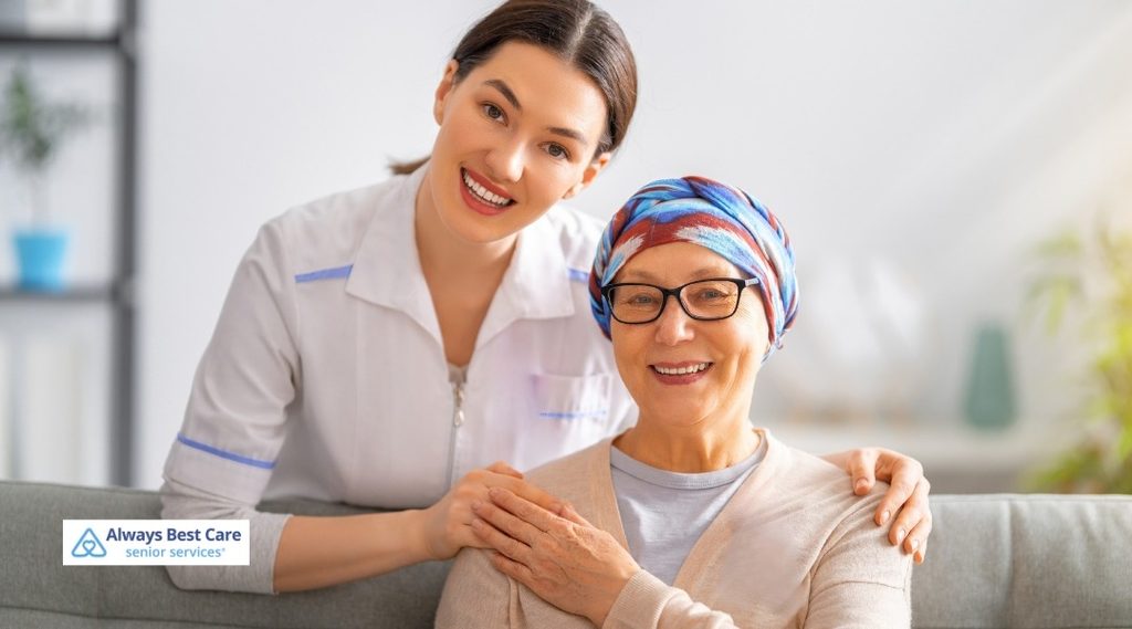 Caregiver resting her hands on the shoulders of a smiling older woman wearing glasses and a colorful headscarf.
