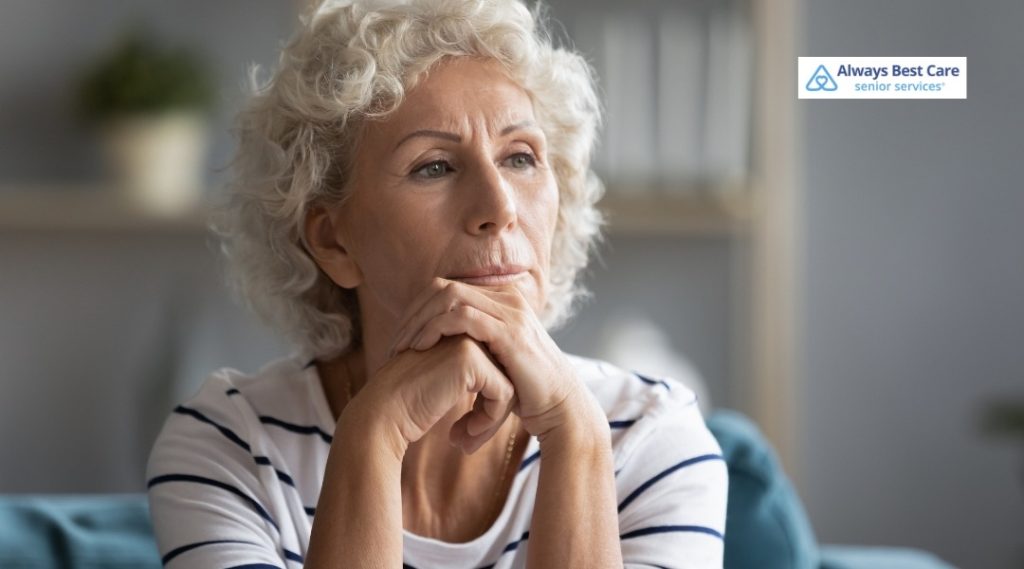 ELDER ABUSE PREVENTION: PROTECTING SENIORS AND PROMOTING SAFE, RESPECTFUL CARE IN HOLLYWOOD, FL 4 OLDER WOMAN SITTING WITH HANDS FOLDED UNDER HER CHIN, LOOKING THOUGHTFUL OR CONCERNED IN A LIVING ROOM, ALWAYS BEST CARE LOGO IN CORNER.