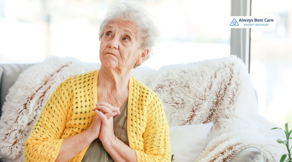 ELDER ABUSE PREVENTION: PROTECTING SENIORS AND PROMOTING SAFE, RESPECTFUL CARE IN HOLLYWOOD, FL 5 OLDER WOMAN SITTING ON A COZY SOFA WITH FLUFFY PILLOWS, HANDS CLASPED NEAR HER CHEST AND WEARING A YELLOW CARDIGAN, ALWAYS BEST CARE LOGO IN CORNER.