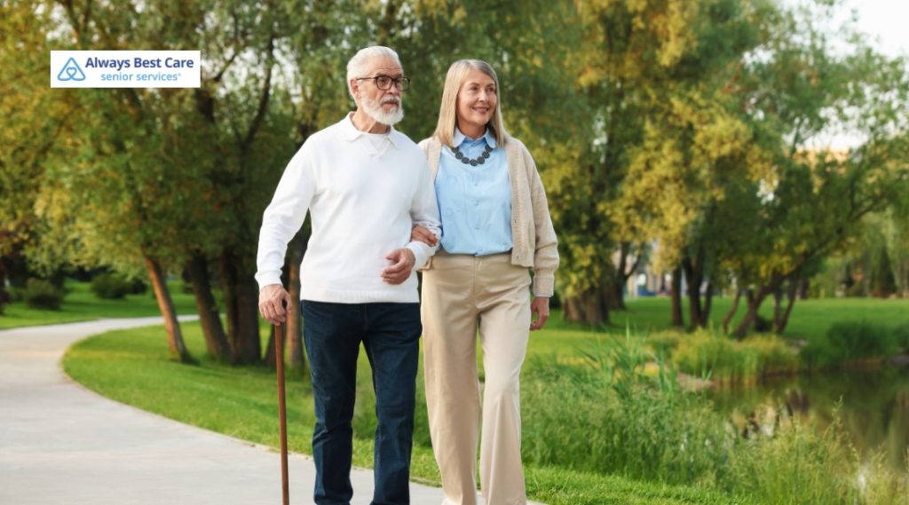 OLDER COUPLE WALKING TOGETHER IN A PARK, WITH THE MAN USING A CANE.