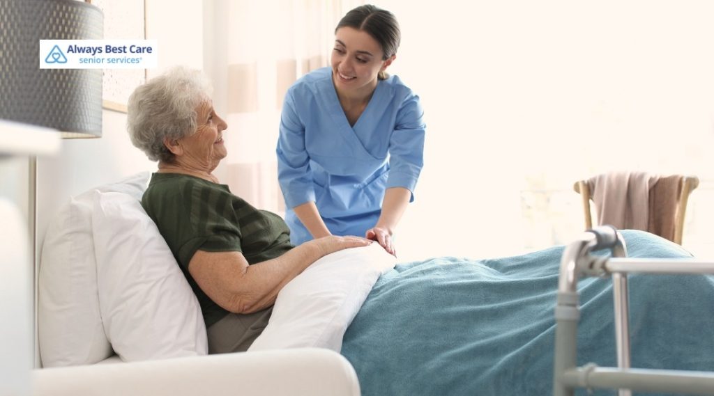 CAREGIVER HOLDS HANDS WITH A SMILING OLDER WOMAN RESTING COMFORTABLY IN BED.