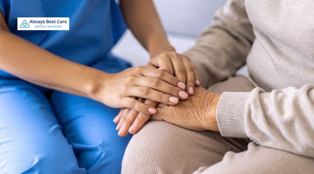 CAREGIVER IN BLUE SCRUBS GENTLY HOLDS AN OLDER ADULT’S HANDS.