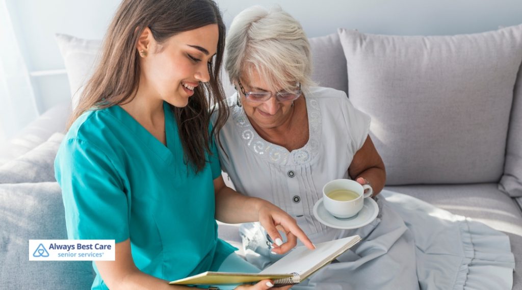 A caregiver dressed in teal scrubs sits beside an elderly woman, both engaged in reading a book together, with the elderly woman holding a cup of tea.