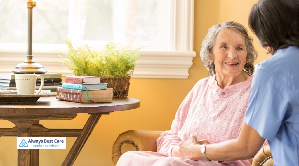 An elderly woman in a pink sweater smiles warmly while holding hands with a caregiver in a blue uniform, seated comfortably in a sunlit living room with books and a coffee mug on a nearby table.