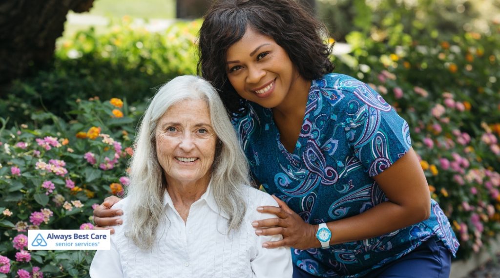 A senior woman with long gray hair and a white blouse smiles while sitting outdoors next to a caregiver in a colorful blue uniform, both beaming and surrounded by blooming flowers.