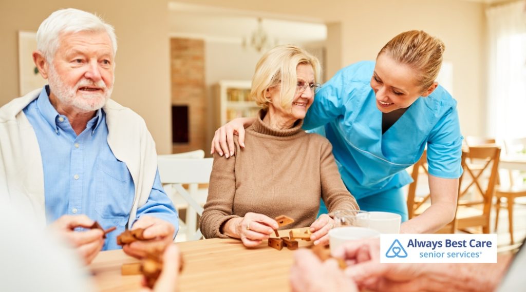 A caregiver interacts with three seniors at a table, helping them with a game. The scene captures a lively moment of companionship and care, with the elderly individuals engaged and enjoying their time together.