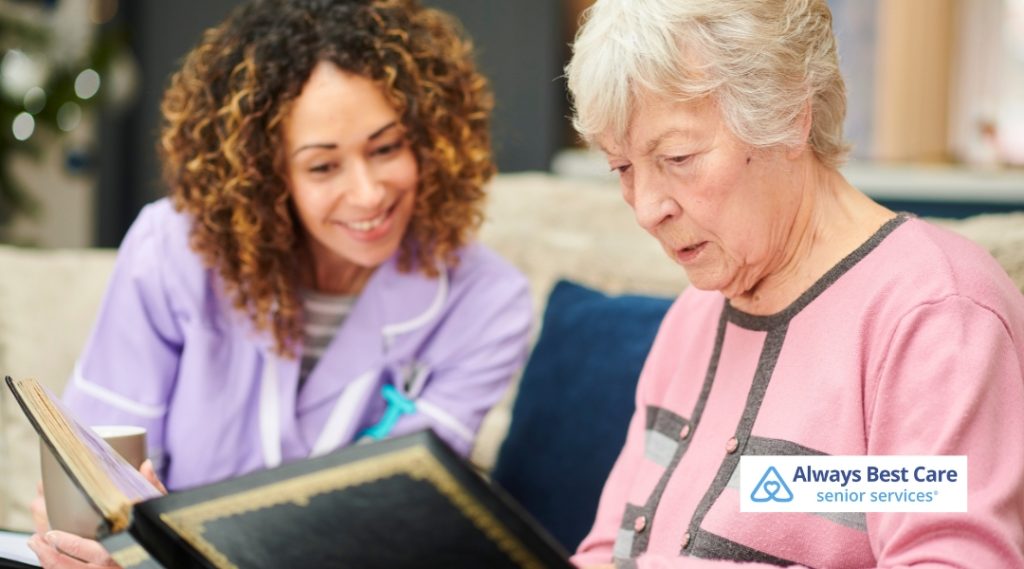 A caregiver and an elderly woman look at a photo album together. The caregiver smiles as they share a peaceful and engaging moment, with the senior woman focusing on the album, immersed in the memories.