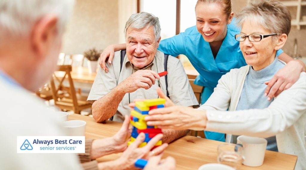 Seniors sit together around a table, smiling and playing a stacking game with colorful blocks. A caregiver assists, creating a warm and engaging atmosphere, where seniors enjoy both fun and social interaction.