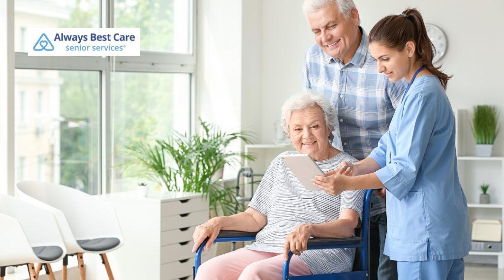 A caregiver in a blue uniform is helping an elderly woman in a wheelchair. The caregiver is showing something on a tablet to the elderly woman, while a man stands beside them, smiling at the screen.