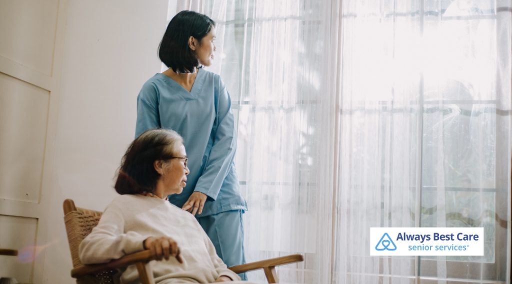 A caregiver in a blue uniform is standing next to an elderly person seated in a chair, looking out the window. The scene conveys a sense of calm and support as the caregiver offers assistance.