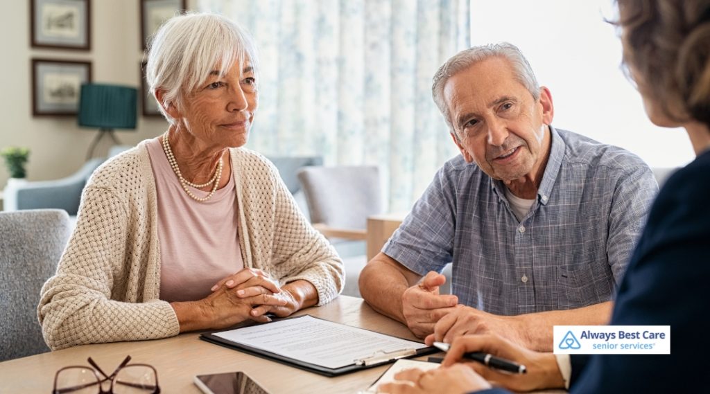 A senior man and woman sit together in a cozy home setting, attentively listening to a caregiver who is speaking with them across the table. A clipboard and pen are on the table, indicating a care consultation.