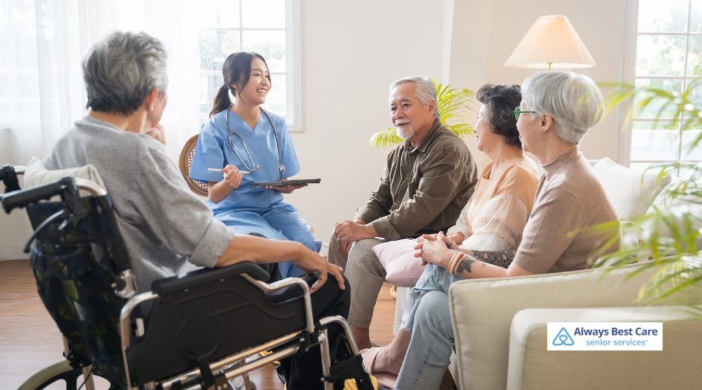 A group of seniors and a caregiver gather in a bright, inviting living room. The caregiver, dressed in medical scrubs, holds a clipboard and engages warmly with the group, representing social and medical support.