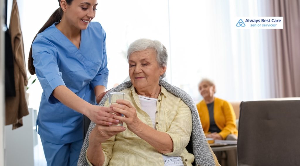 A smiling caregiver hands a glass of water to an elderly woman sitting comfortably with a blanket around her shoulders. Another senior is seen in the background, emphasizing attentive, personal care.