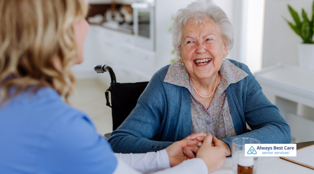 A cheerful elderly woman in a wheelchair holds hands with a caregiver at a kitchen table. They are smiling at each other, highlighting companionship and compassionate support.