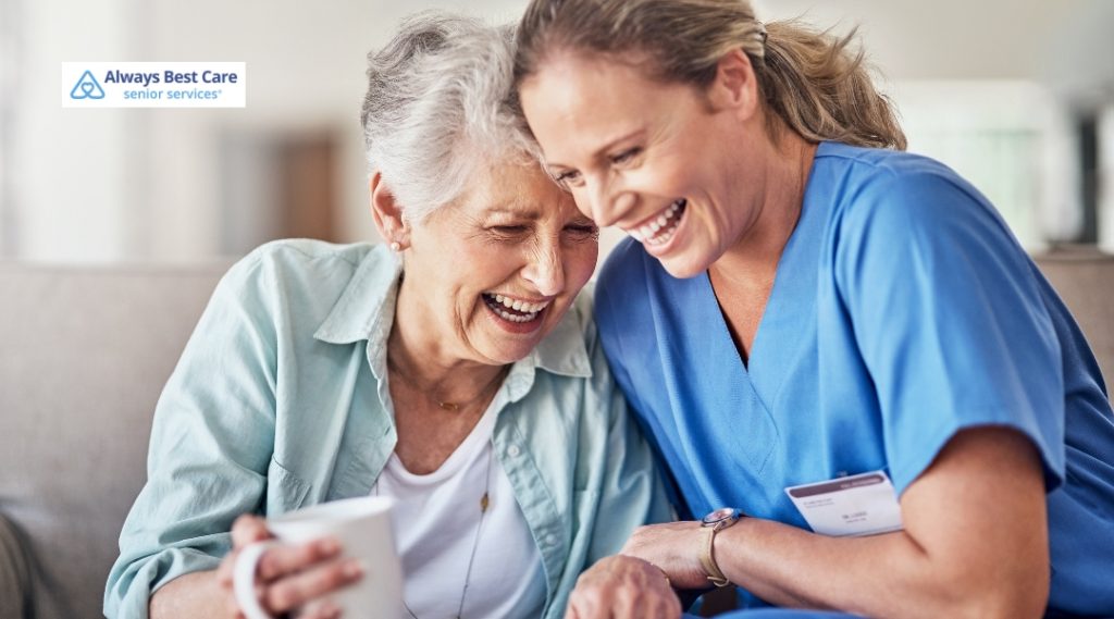 An elderly woman and her caregiver laugh together while sitting closely on a couch. Both appear joyful and relaxed, symbolizing a strong caregiver-client bond.