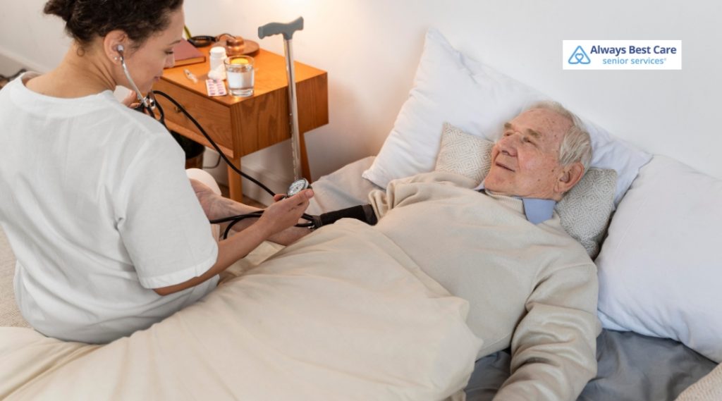 A smiling female caregiver in blue scrubs gently holds hands with an elderly woman lying comfortably in bed. They share a warm, caring moment in a bright, sunlit bedroom. The Always Best Care logo is visible in the top right corner.