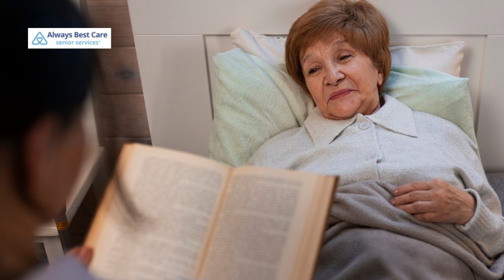 An elderly woman in a light gray coat rests in bed, looking attentively at a caregiver who is reading a book to her. The scene conveys a peaceful and compassionate home care environment. The Always Best Care logo is in the top left corner.