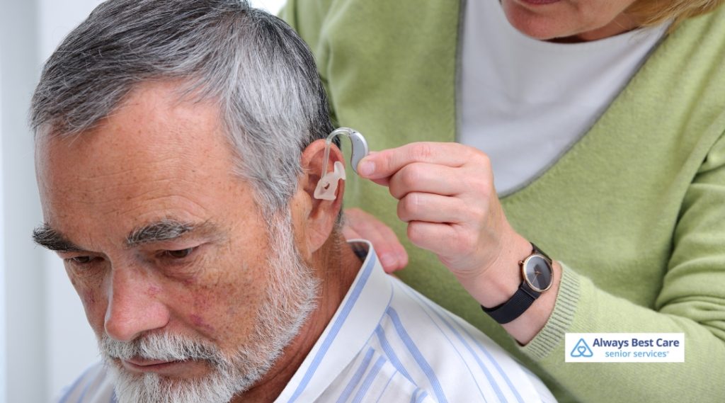A caregiver helps a senior man by placing a hearing aid in his ear in a healthcare setting.
