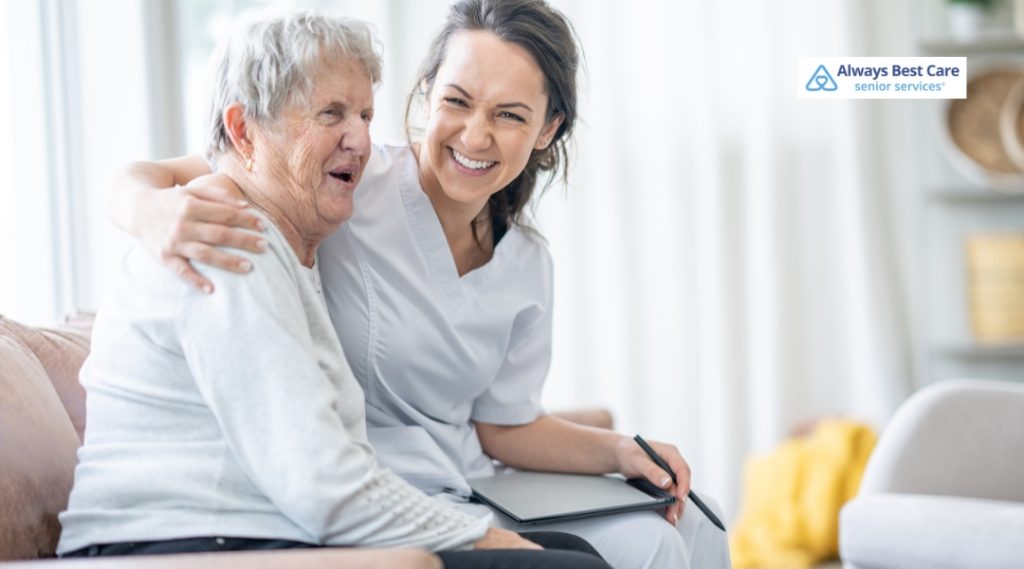 A caregiver and elderly woman sharing a joyful moment, sitting together with smiles on their faces, in a comfortable living room.