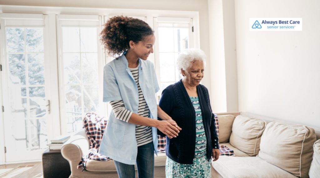 A caregiver assisting an elderly woman with walking in a bright, airy living room, with the caregiver supporting her arm.