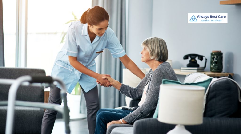A caregiver gently holding the hand of an elderly woman, offering assistance in a living room setting, with natural light streaming in.
