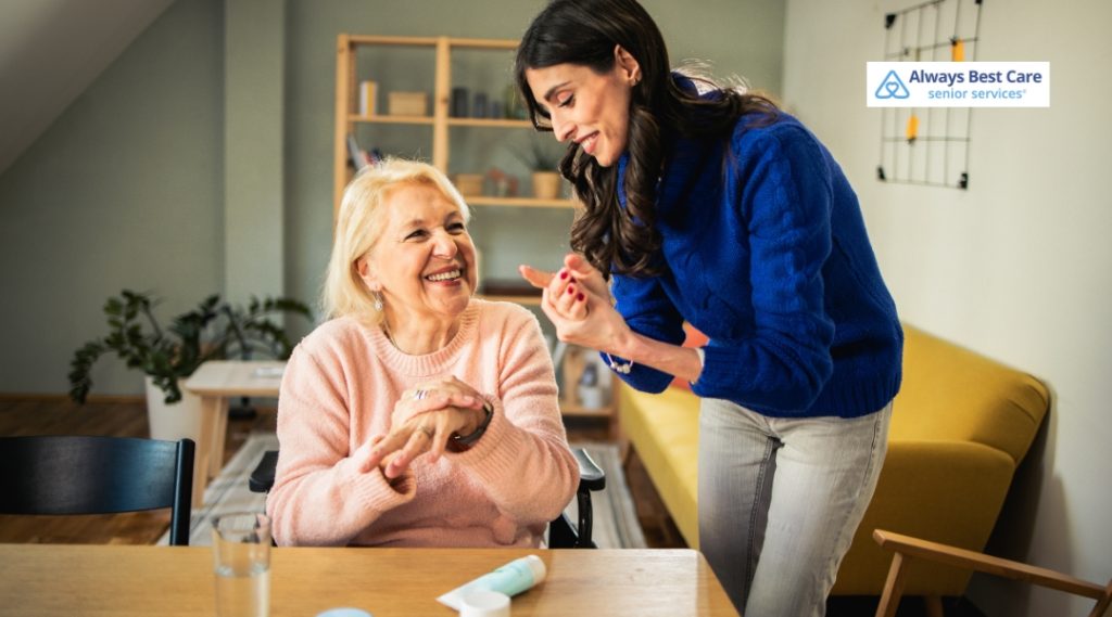 A caregiver smiling while applying cream to the hands of a senior woman in a pink sweater. The woman is sitting in a wheelchair and looking joyful as she enjoys the care provided by the caregiver, who is wearing a blue sweater. The image features Always Best Care’s logo.
