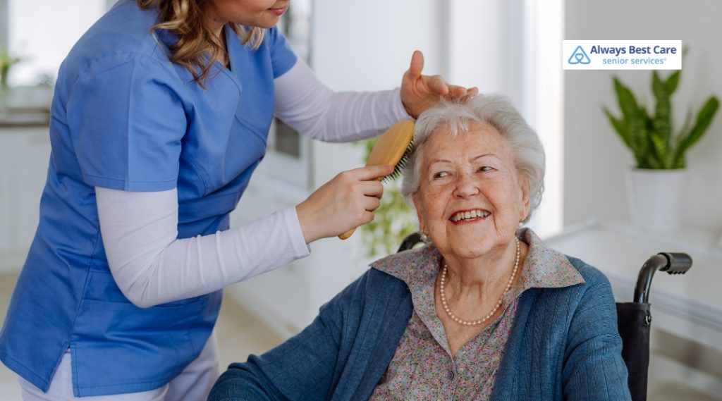 A caregiver, dressed in blue scrubs, brushing the hair of a smiling senior woman with gray hair in a wheelchair. The senior woman looks pleased as the caregiver takes care of her grooming. The Always Best Care logo is in the corner.