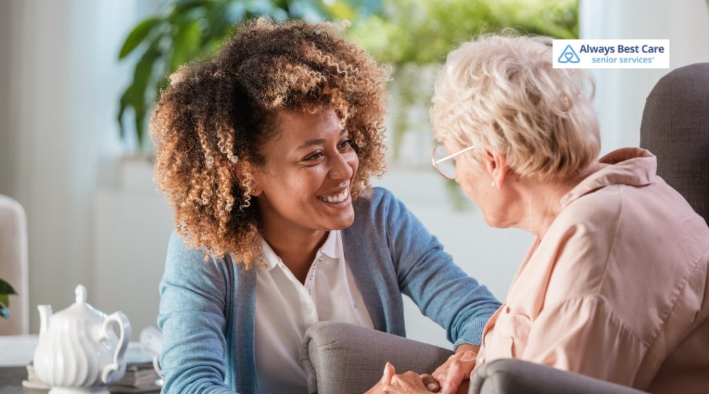 A caregiver with curly hair, wearing a blue cardigan, talking to a senior woman sitting in a chair. The caregiver is smiling warmly while holding the senior's hands, and the senior woman is also smiling, enjoying the conversation. The Always Best Care logo appears in the top corner.