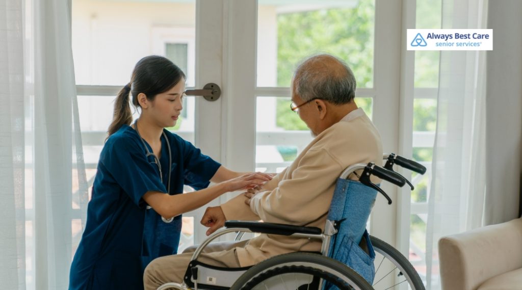 A caregiver in blue scrubs is assisting an elderly man in a wheelchair, gently adjusting his arm while in a well-lit room with large windows.