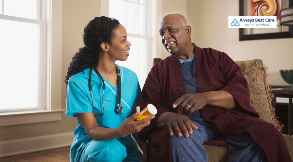 A caregiver with a stethoscope is sitting with an elderly man, holding a bottle of medication and smiling. They are both sitting in a cozy, well-lit living room.