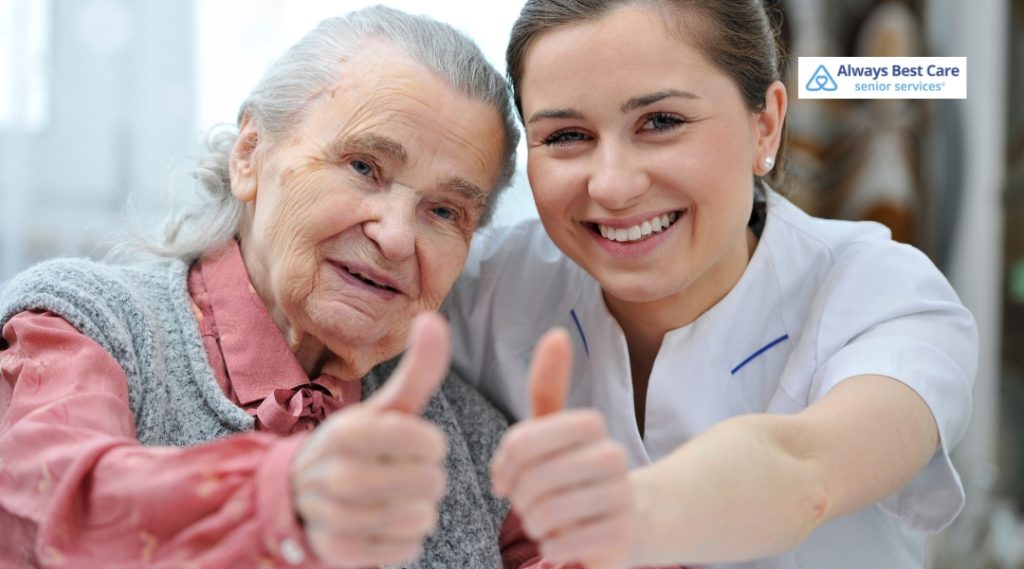 A caregiver and an elderly woman are giving a thumbs-up in a bright and cheerful environment. Both are smiling, with the caregiver wearing a white uniform and the elderly woman in a pink blouse.