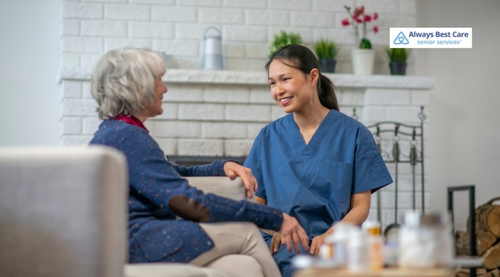 A caregiver is talking to an elderly woman while sitting on a sofa in a comfortable living room. The elderly woman is smiling, and they both seem engaged in conversation.