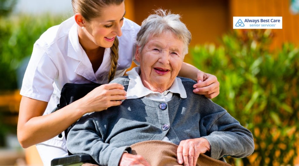 A caregiver is assisting an elderly woman in a wheelchair, helping her by adjusting her sweater. They are outside, with greenery and bright sunlight around them. The caregiver is wearing a white uniform, and the elderly woman has a gray sweater.