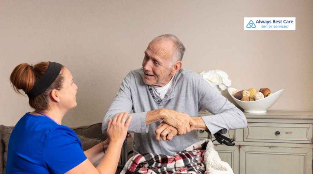 A caregiver is assisting an elderly man in a wheelchair, holding his hand gently. The elderly man is smiling and wearing a gray shirt, while the caregiver, dressed in blue scrubs, is offering support in a cozy indoor setting.