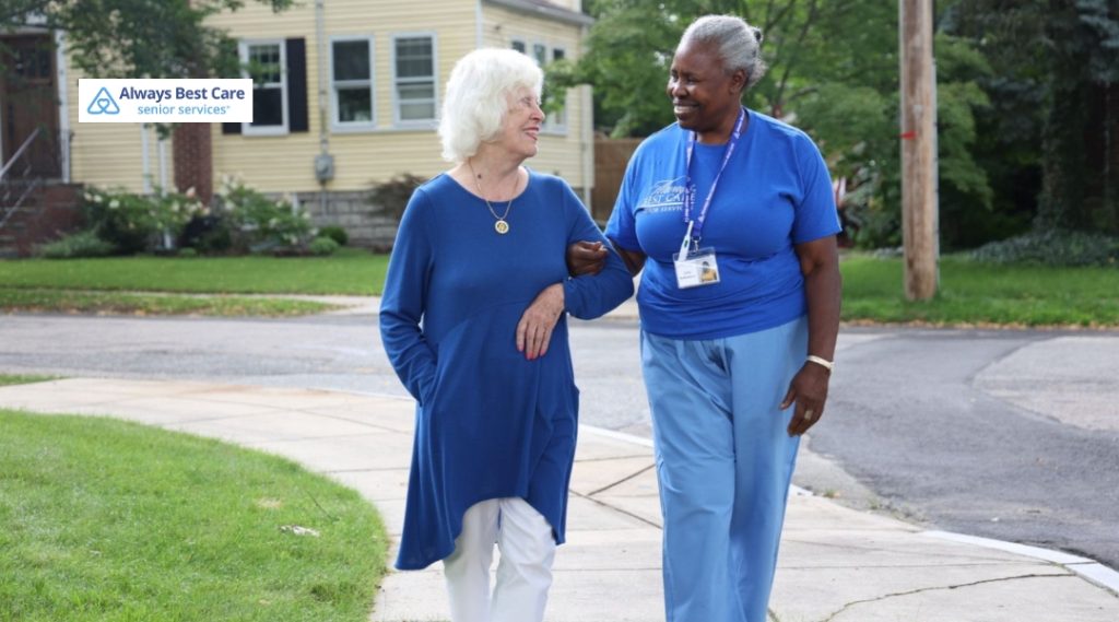 Caregiver walking arm-in-arm with an older woman along a neighborhood sidewalk, smiling at each other.