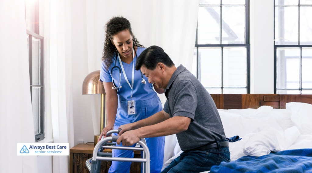A nurse in blue scrubs is assisting a man with a walker as he rises from his bed in a sunlit room, focusing on ambulatory support and rehabilitation.