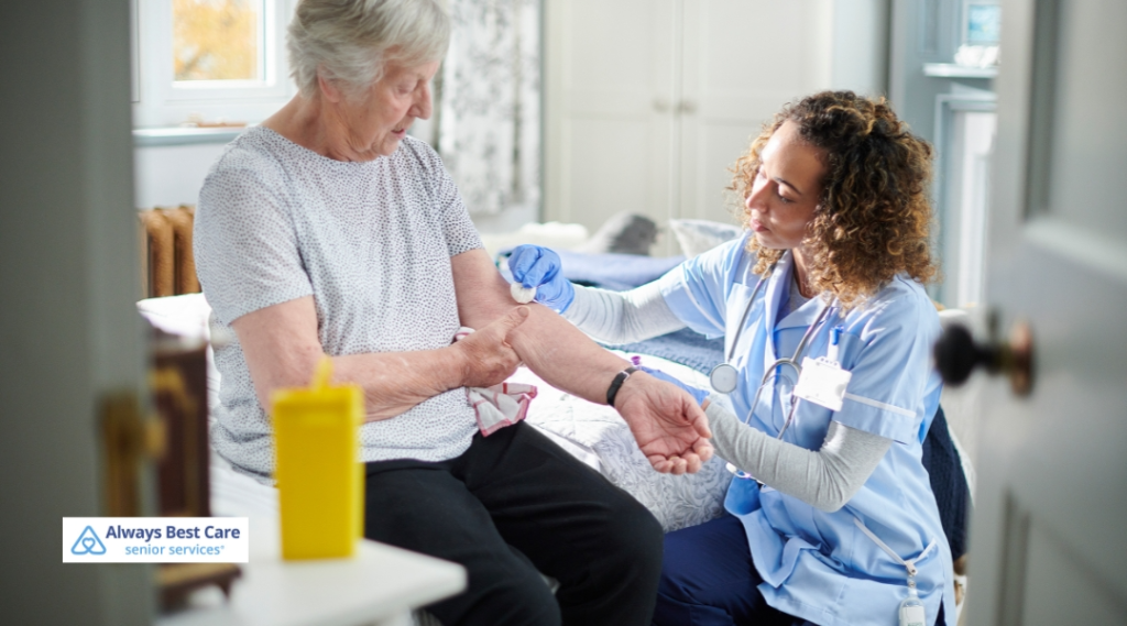 A nurse in blue scrubs is gently preparing to administer an injection or take a blood sample from the arm of an elderly woman seated on a bed in a well-lit bedroom. 