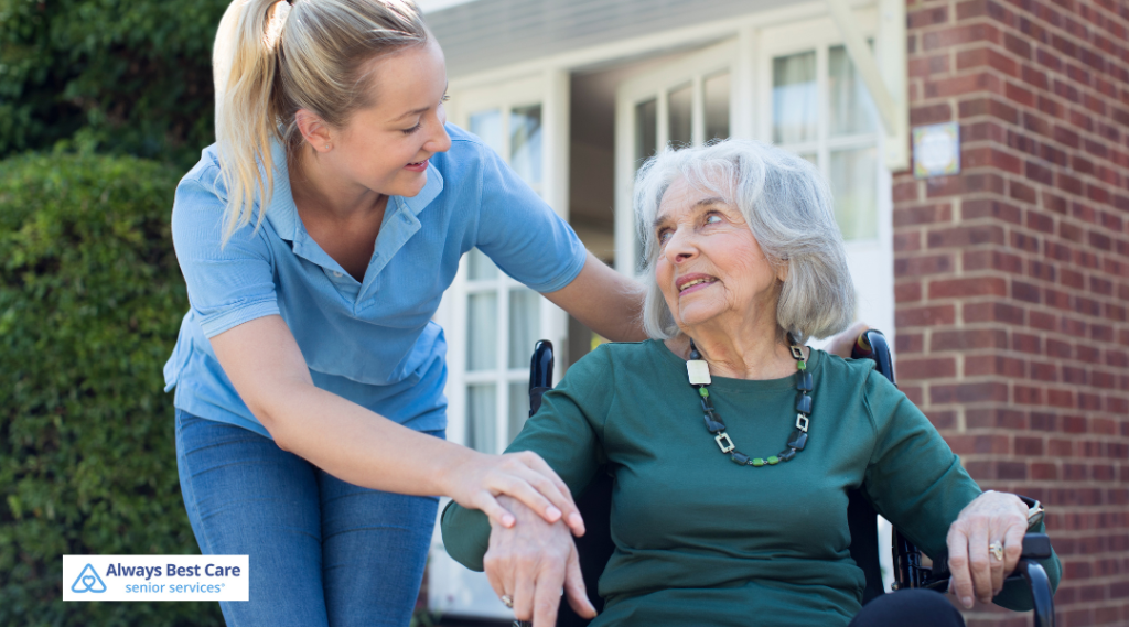 An elderly woman sits outside in a wheelchair, reading a book, while a male caregiver in blue scrubs kneels nearby holding a coffee cup and attentively engages with her.