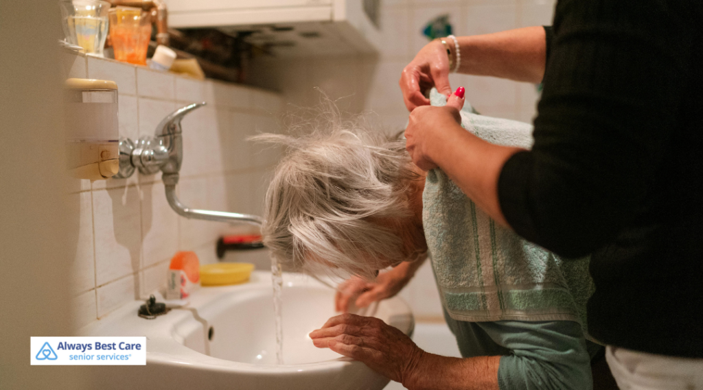  A caregiver assists an elderly woman with washing her hair at a bathroom sink while wearing a towel around her shoulders. 