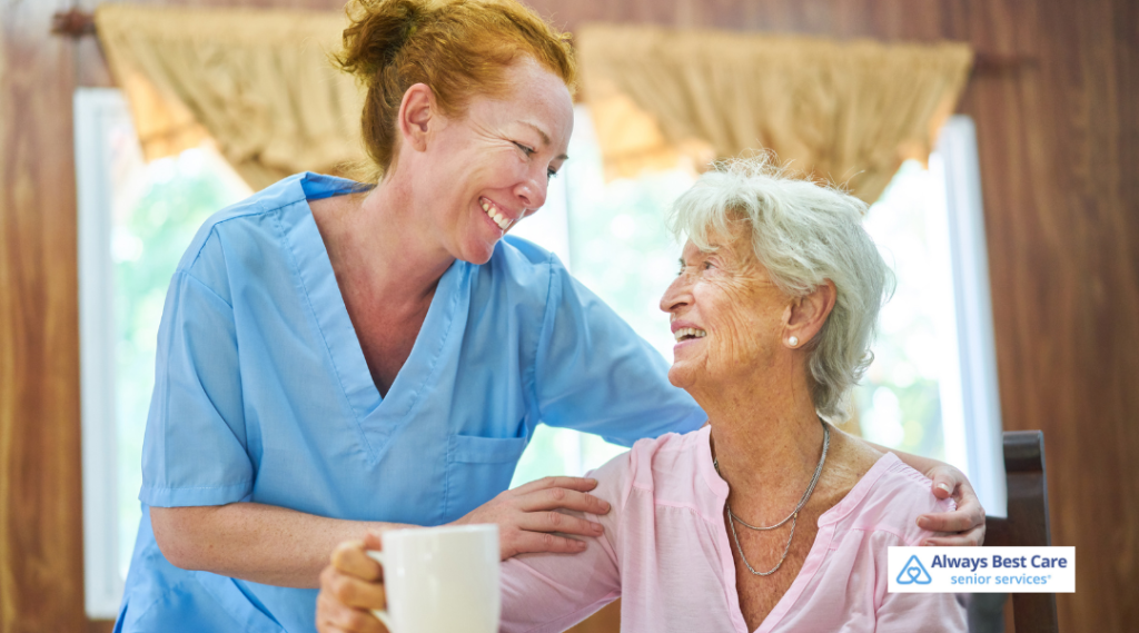 A caregiver in light blue scrubs gently places a supportive hand on the shoulder of a smiling senior woman seated indoors, with a cup in hand. 