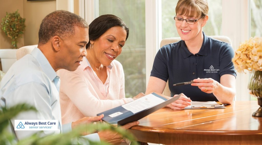 A professional Always Best Care representative, dressed in a navy polo shirt, sits at a table and discusses paperwork with a couple.