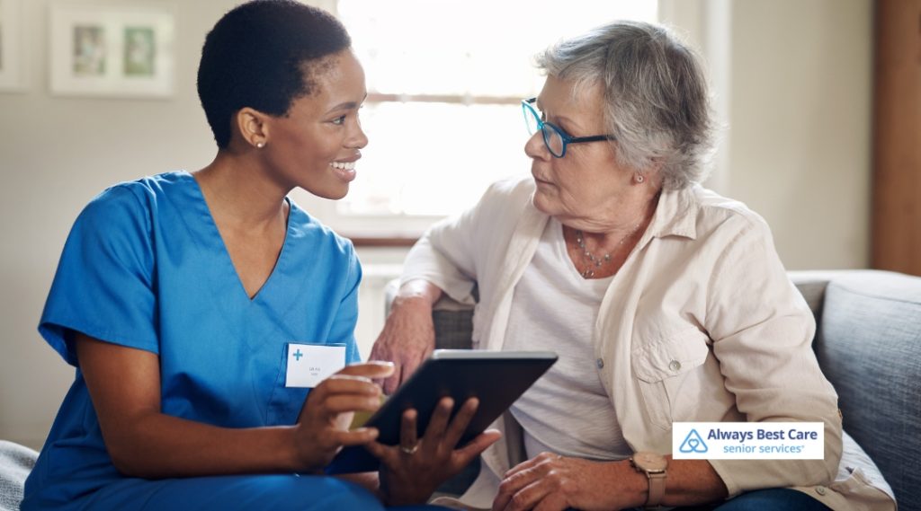 A female caregiver in blue scrubs sits with an older woman on a couch, holding a tablet and discussing care options. The Always Best Care Senior Services logo is visible in the bottom right corner.