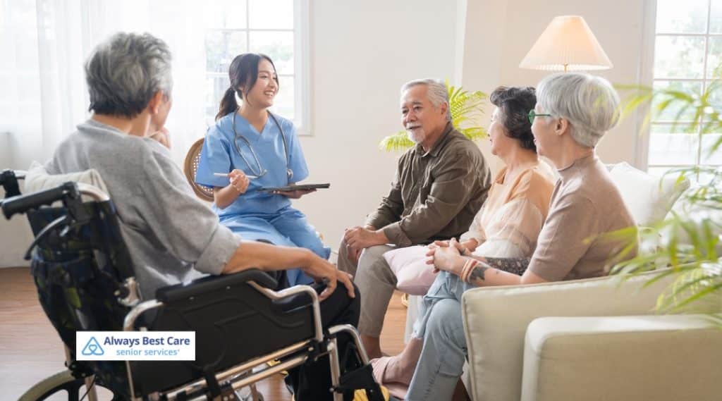 A female caregiver in light blue scrubs sits facing two older adults, speaking animatedly and referencing notes in an open binder. The Always Best Care Senior Services logo is present in the lower left corner.
