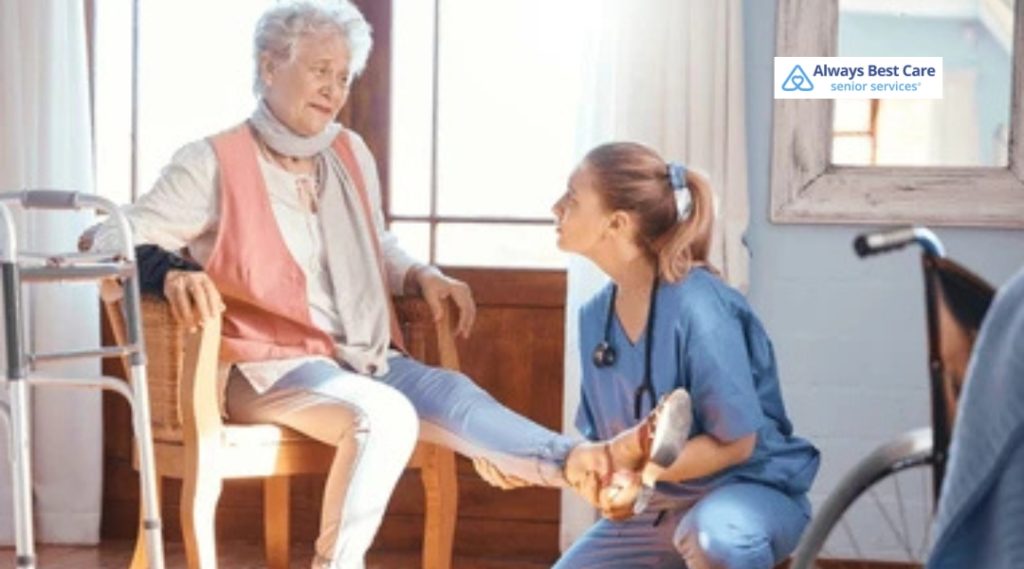 A caregiver assists an elderly woman with her leg, possibly helping with a mobility or rehabilitation exercise in a home setting.
