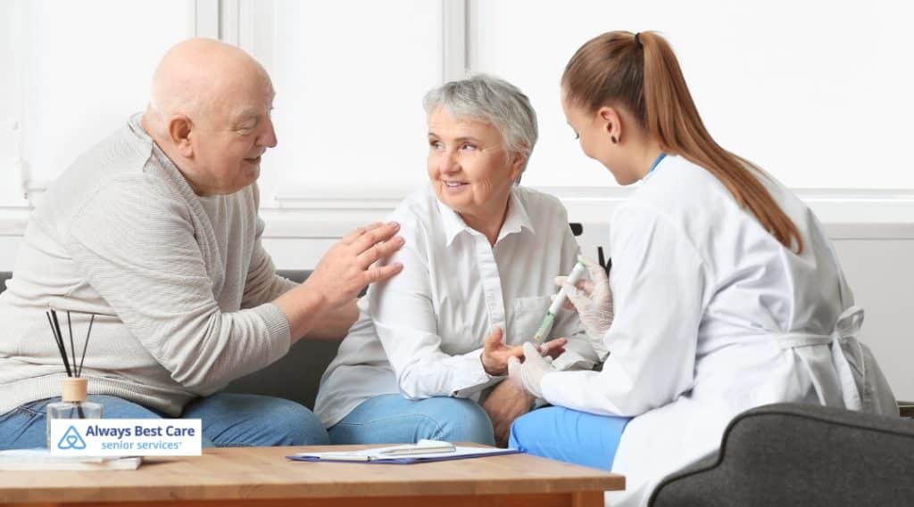 A caregiver and a senior couple reviewing diabetes management, with the caregiver explaining the importance of medication and blood glucose monitoring.